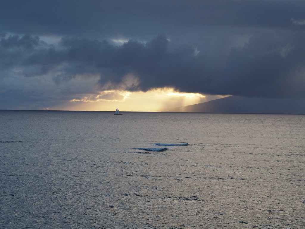 A cloudy sunset, featuring a sailboat near Molok'ai, shot from a balcony in Kahana